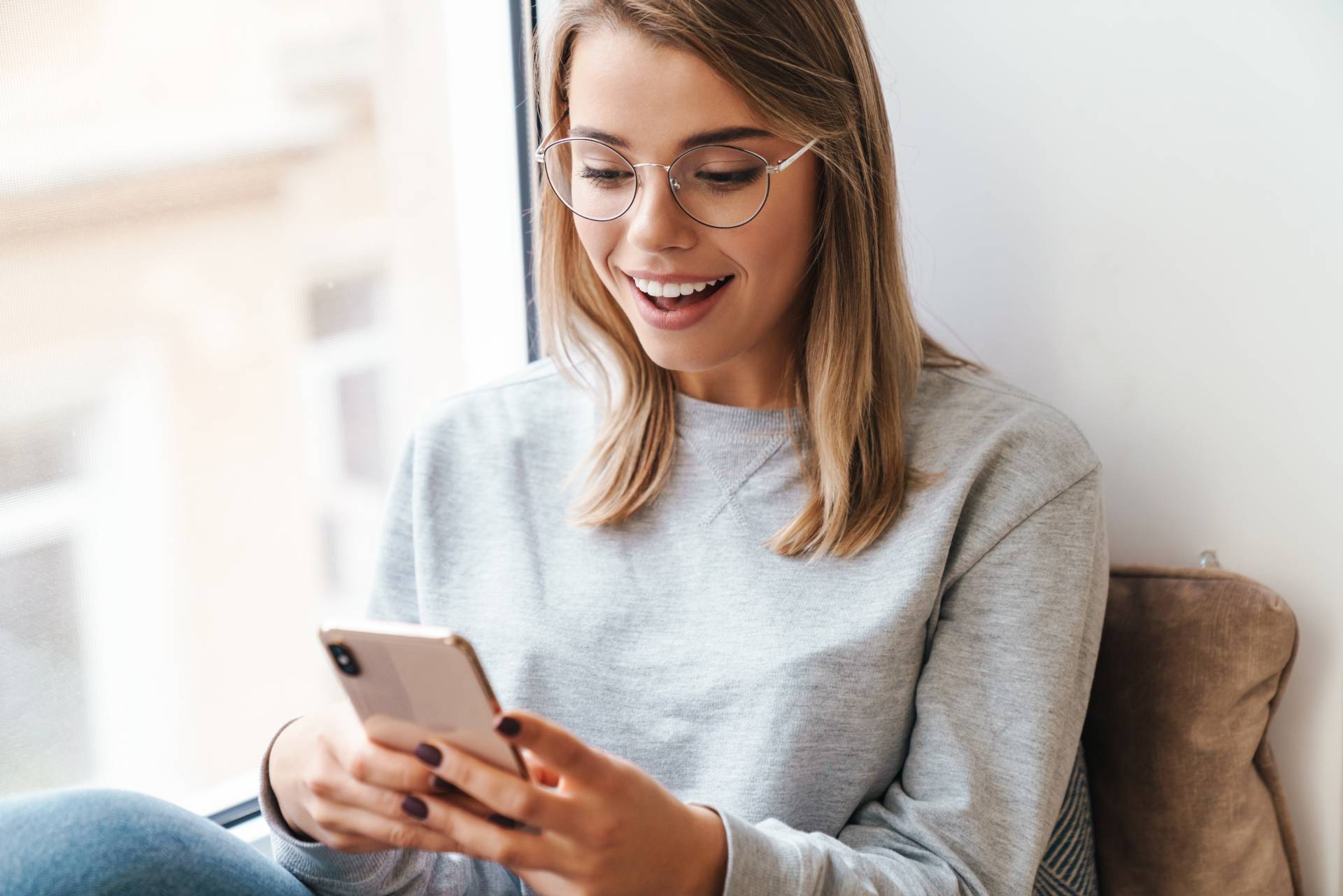 A happy woman using a mobile phone while seated by a glass window