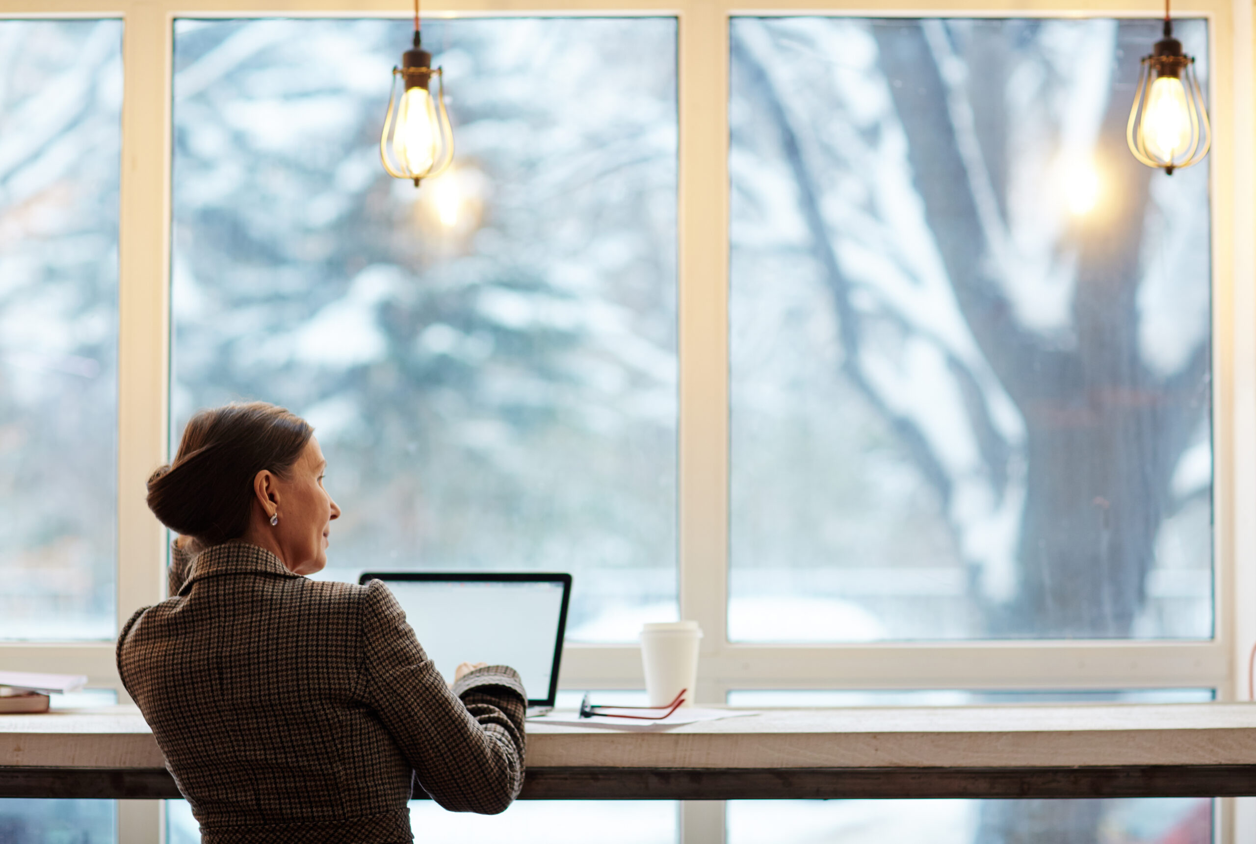 Woman looking out the window while seated by a long table working on a laptop